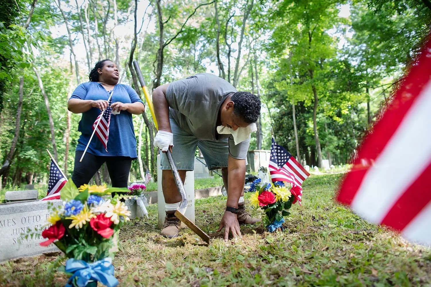 Two volunteers tending graves in a wooded cemetery, with flags and flowers beside the headstones